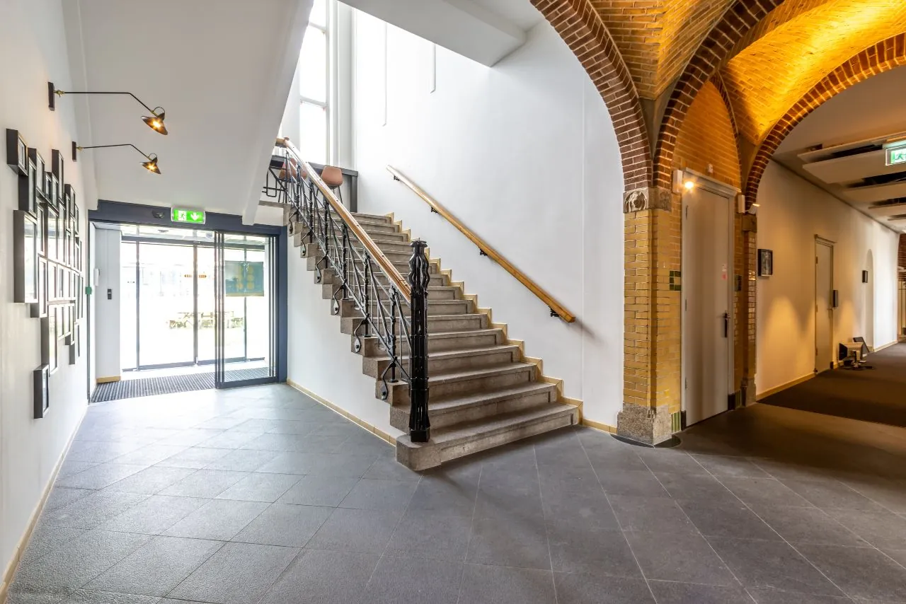 Ingangshal met een stenen trap, gewelfde bakstenen plafonds en glazen schuifdeuren in een gebouw aan de Lage Naarderweg.